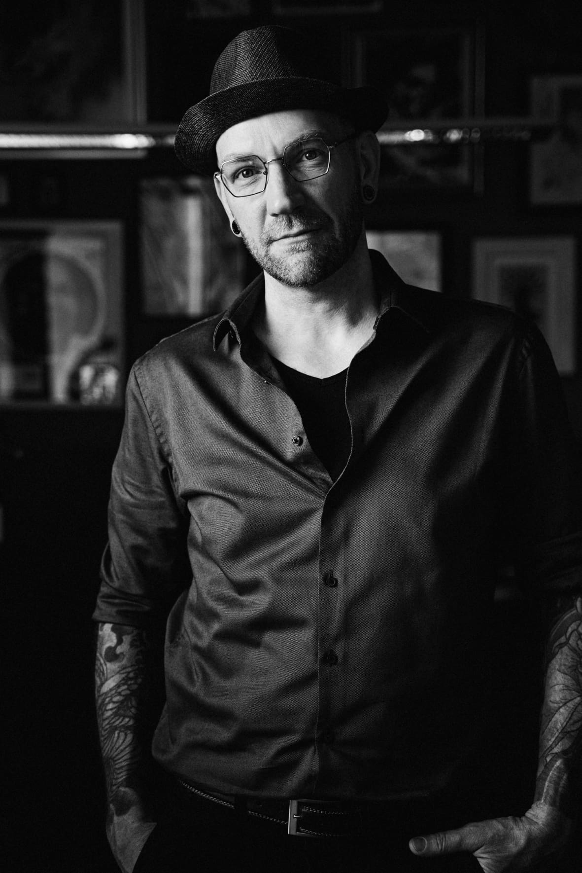 Black-and-white portrait of a man wearing a hat and glasses, with a short beard, in a dimly lit room; visible tattoos on his forearms.